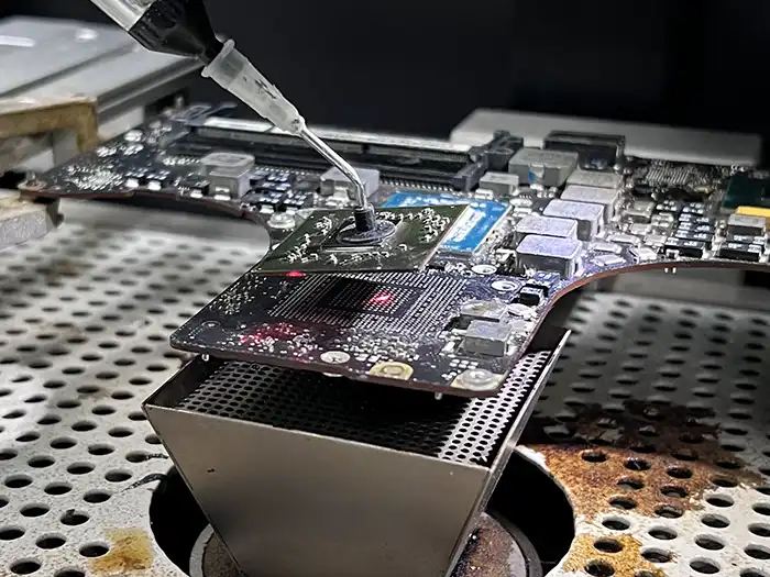 A MacBook logic board on a preheater with flux applied to a chip site during rework