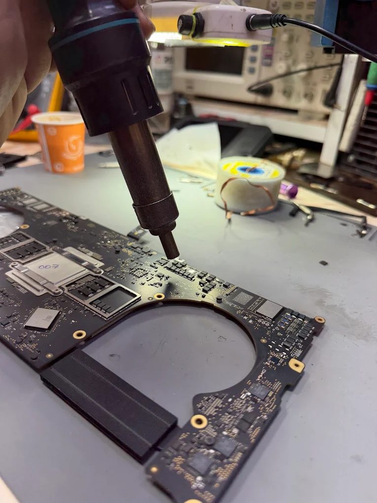 A hot-air nozzle held over components on a MacBook Pro logic board under a bench light